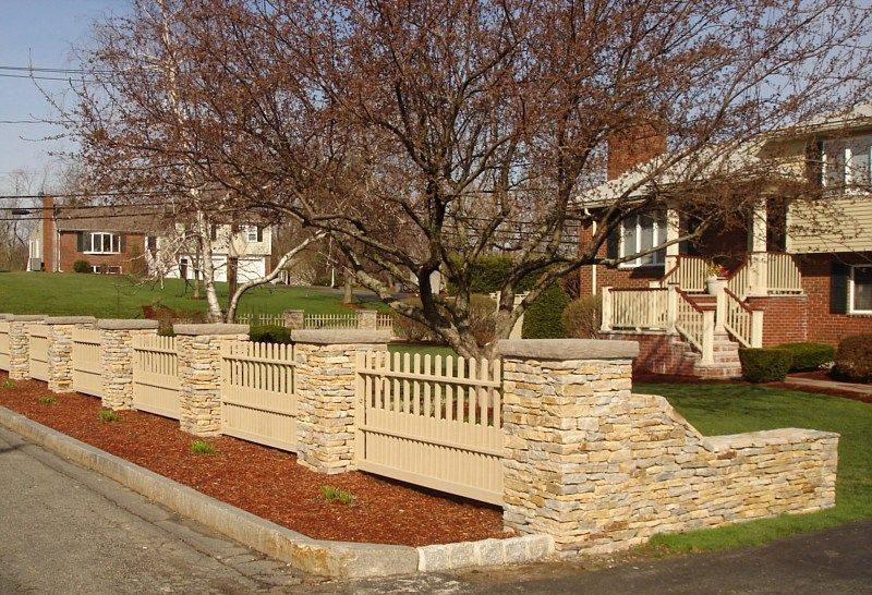 A stone fence with tan wooden slats borders a garden bed of red mulch in front of a suburban brick home.