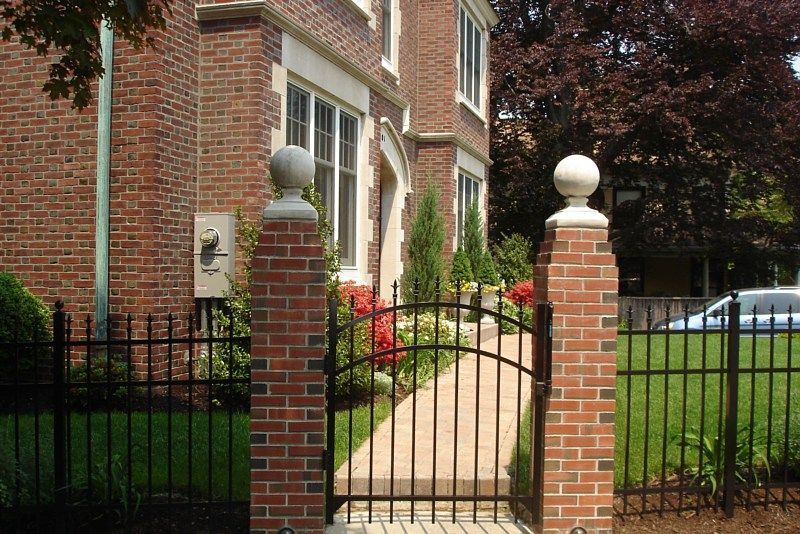 A black metal gate and fence set between two brick pillars topped with stone spheres in front of a brick house.
