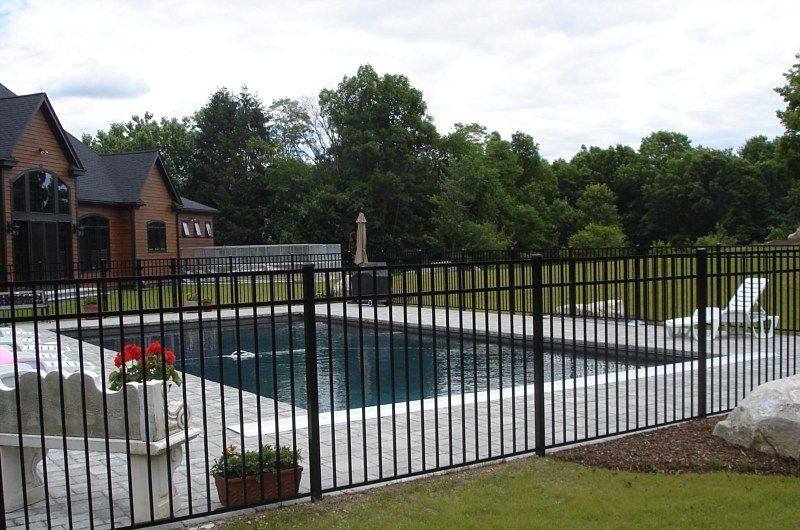 A rectangular backyard swimming pool surrounded by a black metal fence, a stone patio, and a house in the background.