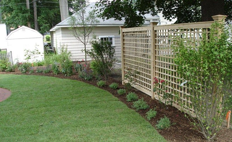 A wooden lattice garden fence stands in a yard, bordered by a fresh mulch bed with small plants and a curved green lawn.