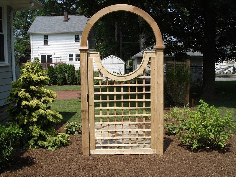 A wooden garden gate with a circular top cutout, positioned in a yard between two bushes with a house in the background.