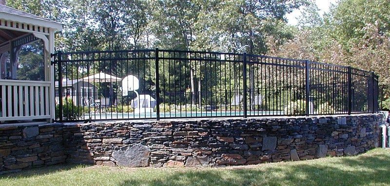 A black metal fence encloses a swimming pool built into a raised, natural stone retaining wall in a backyard.