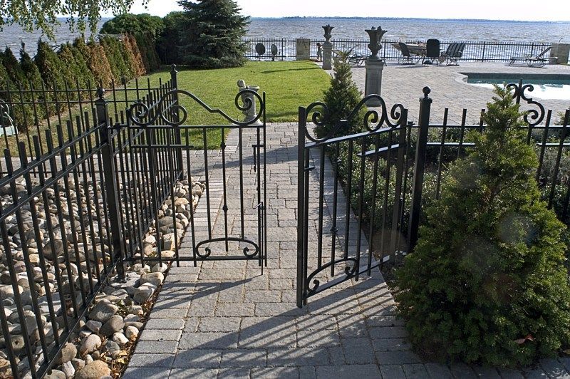 Open black decorative iron gate leading to a paved patio, swimming pool, and lake view.