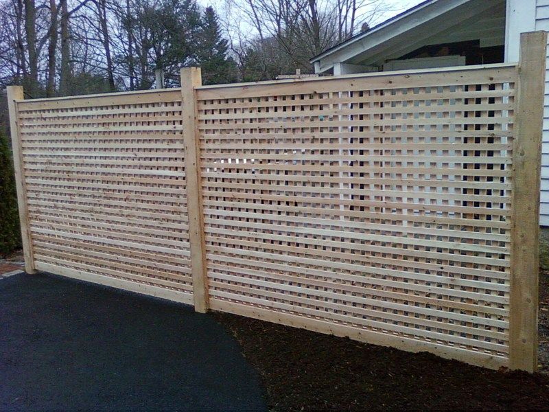 A light wood lattice privacy fence standing next to a house with a paved driveway and mulch bed in the foreground.