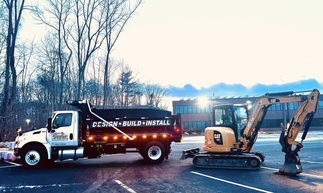 A yellow construction excavator parked next to a white dump truck in an outdoor lot with trees in the background.
