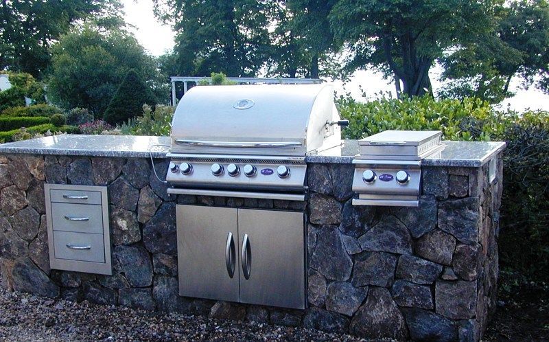 A stainless steel outdoor grill and side burner set into a rustic stone counter in a garden setting.