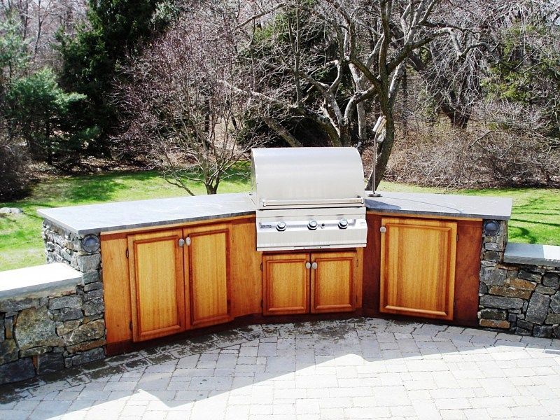 An outdoor kitchen island with wooden cabinets and a stainless steel grill set against a stone wall on a paved patio.