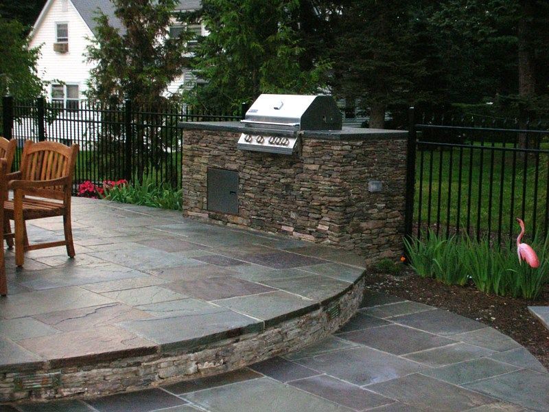 A stone outdoor kitchen with a grill on a raised patio, next to a wooden chair, a black metal fence, and garden plants.