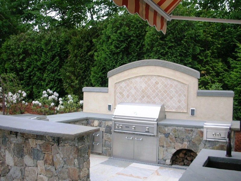 Outdoor kitchen with a stone base, built-in stainless steel grill, a tiled backsplash, and a counter, set in a garden.