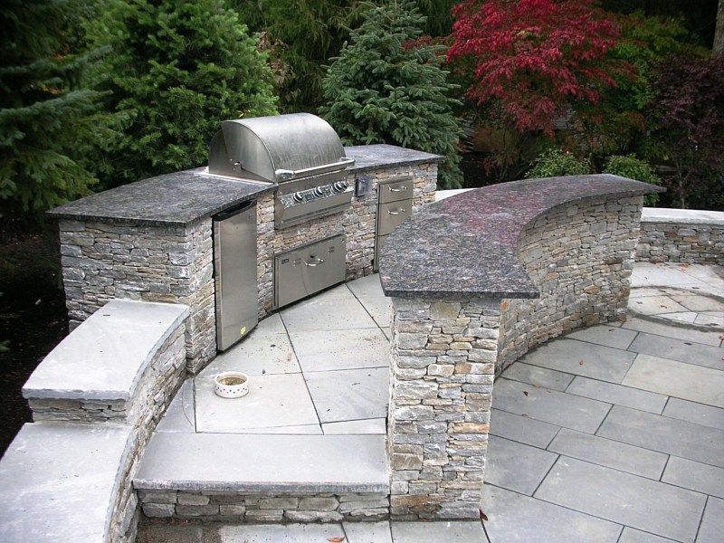 Outdoor kitchen featuring a stone grill station and curved counter on a flagstone patio surrounded by trees and shrubs.