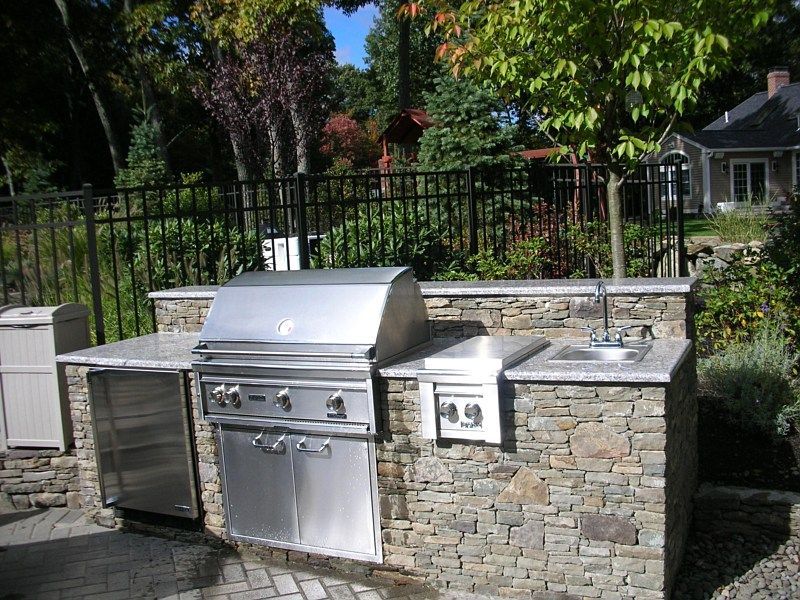 Outdoor stone kitchen island with a stainless steel grill, side burner, sink, and under-counter refrigerator in a backyard.