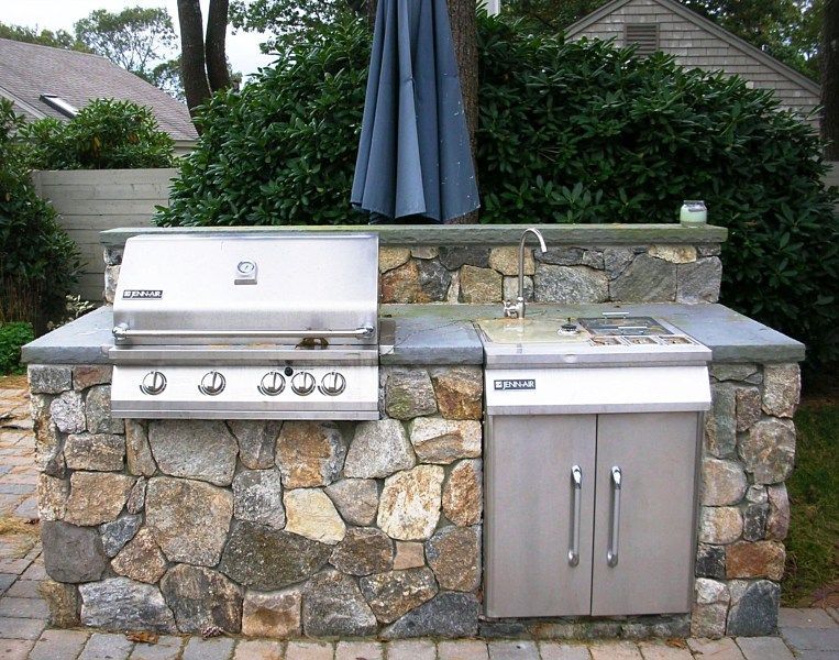 Outdoor stone kitchen island with a stainless steel grill, side burner, and sink, situated in a backyard setting.