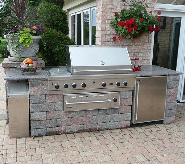 An outdoor kitchen with a stainless steel grill, stone base, and a built-in refrigerator on a stone patio.
