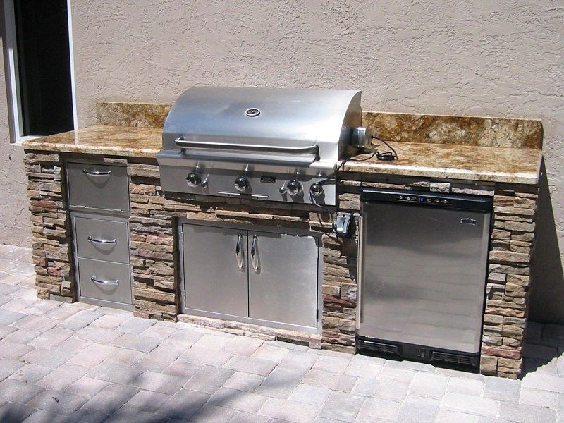 An outdoor kitchen island with stacked stone, a granite countertop, a stainless steel grill, cabinet doors, and a fridge.