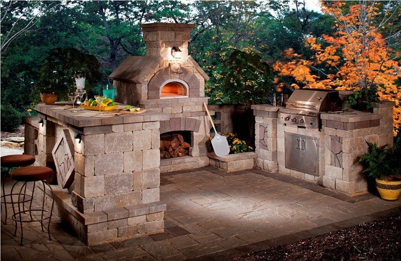 An outdoor kitchen features a stone wood-fired oven, a grill station, and a patio bar area in a wooded setting.