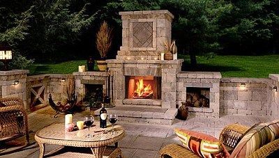 A cozy stone outdoor fireplace at night with a roaring fire, flanked by seating and decorative lighting on a stone patio.