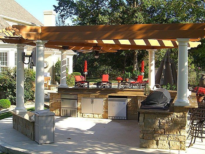 Outdoor kitchen with a wooden pergola, stone counter, grill, and patio seating in a backyard setting.