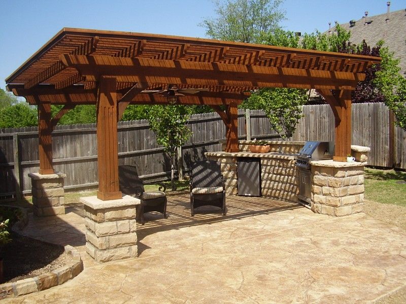 A wooden pergola patio cover with stone pillars, featuring an outdoor kitchen area with a grill on a stamped concrete deck.