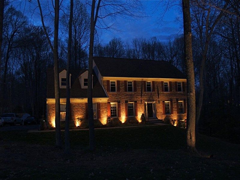 A two-story brick house illuminated by warm exterior up-lighting at dusk, surrounded by dark trees.