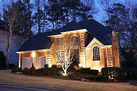 A two-story brick house at dusk, illuminated by warm exterior lights, surrounded by trees and a trimmed lawn.