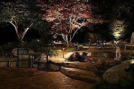 A brick patio illuminated at night, featuring tiered stone steps, garden lighting, and trees under a dark sky.