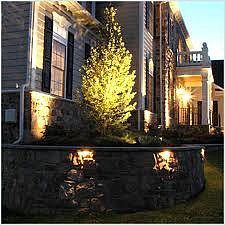 A residential stone retaining wall with integrated path lighting illuminates a glowing tree in front of a house at dusk.