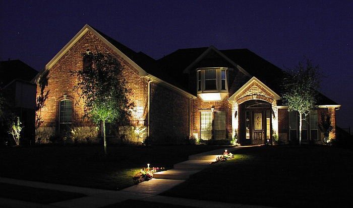 A large brick house at night, illuminated by warm landscape lighting, with a lit walkway leading to the front entrance.