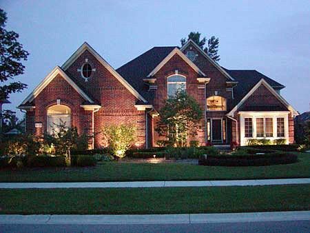 A two-story brick house with a pitched roof and lit windows, surrounded by a lawn and landscaped garden at dusk.