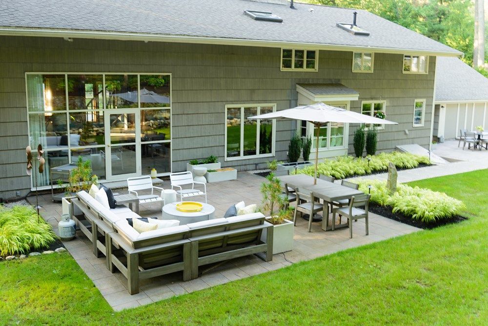A backyard stone patio with a gray L-shaped sectional, a dining set under an umbrella, and surrounding landscaping.