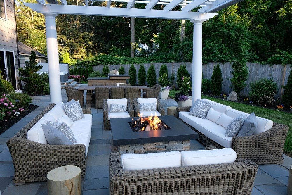 A patio with a fire pit, cushioned wicker sofas, and a white pergola, set in a backyard garden with stone flooring.