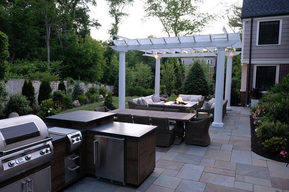 A patio with a stainless steel grill station, a long dining table, and a white pergola covering cushioned outdoor seating.