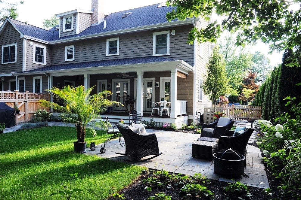 A gray, multi-story house with a covered patio, outdoor wicker furniture, and a grassy lawn on a sunny day.