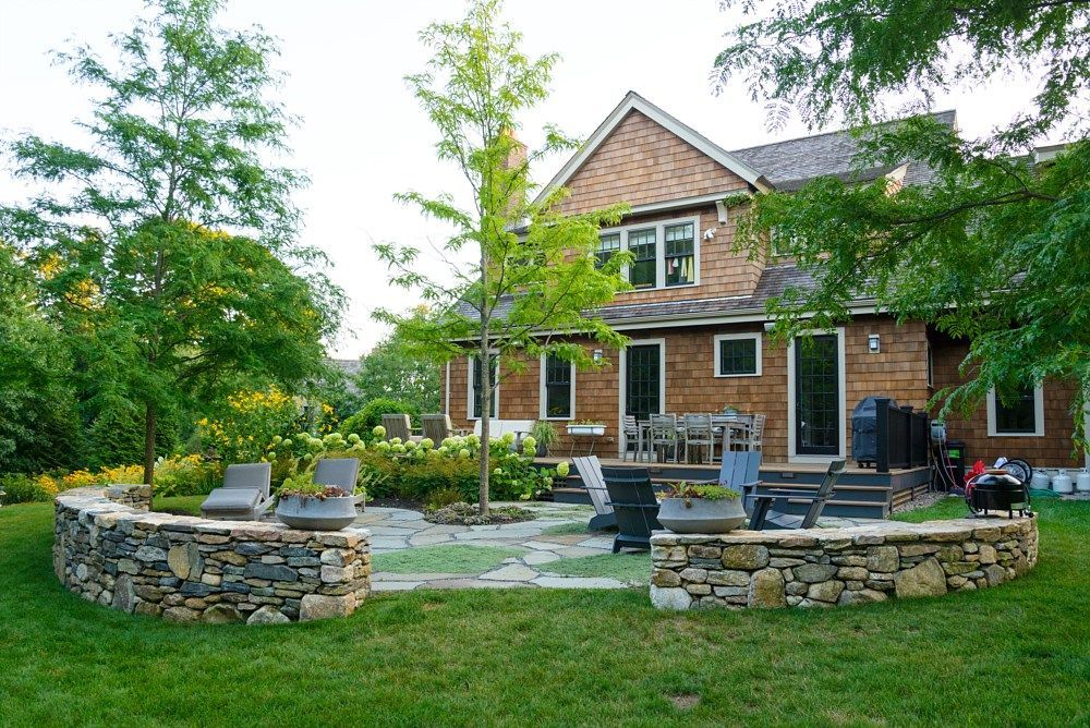 A backyard stone patio with a curved wall, seating, and a central tree in front of a brown shingled house.