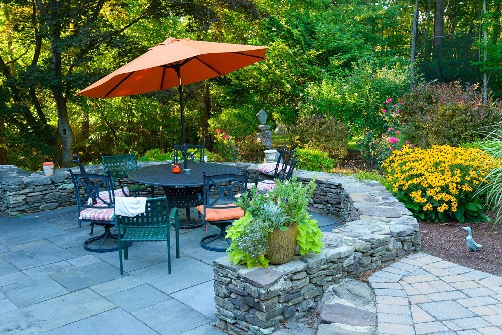 A patio with a round black table, green chairs, and an orange umbrella, surrounded by stone walls and garden flowers.