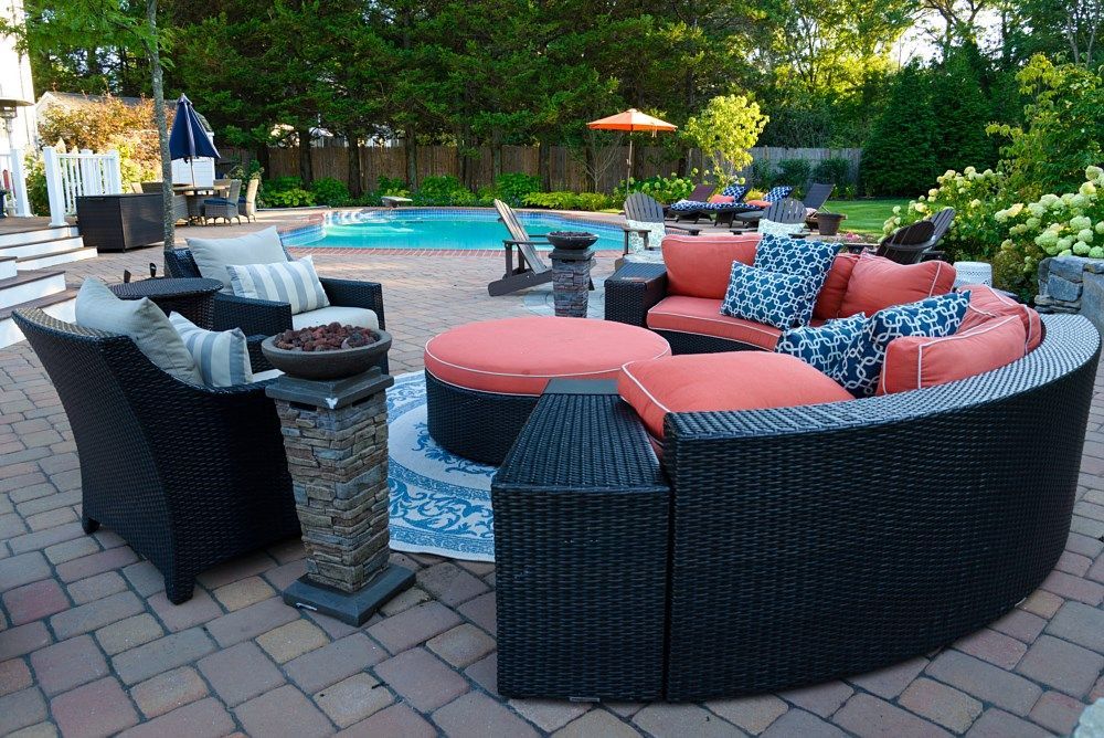 Outdoor patio featuring a curved black wicker sofa with coral cushions and a matching ottoman beside a backyard pool.