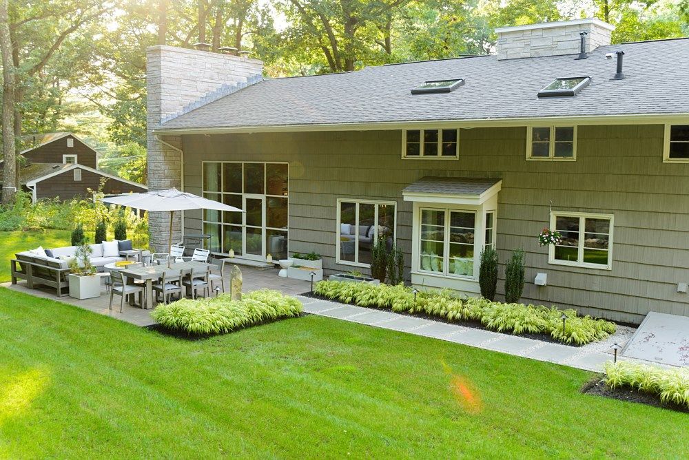 A two-story grey home features a stone chimney, a stone patio with outdoor furniture, and lush green landscaping.