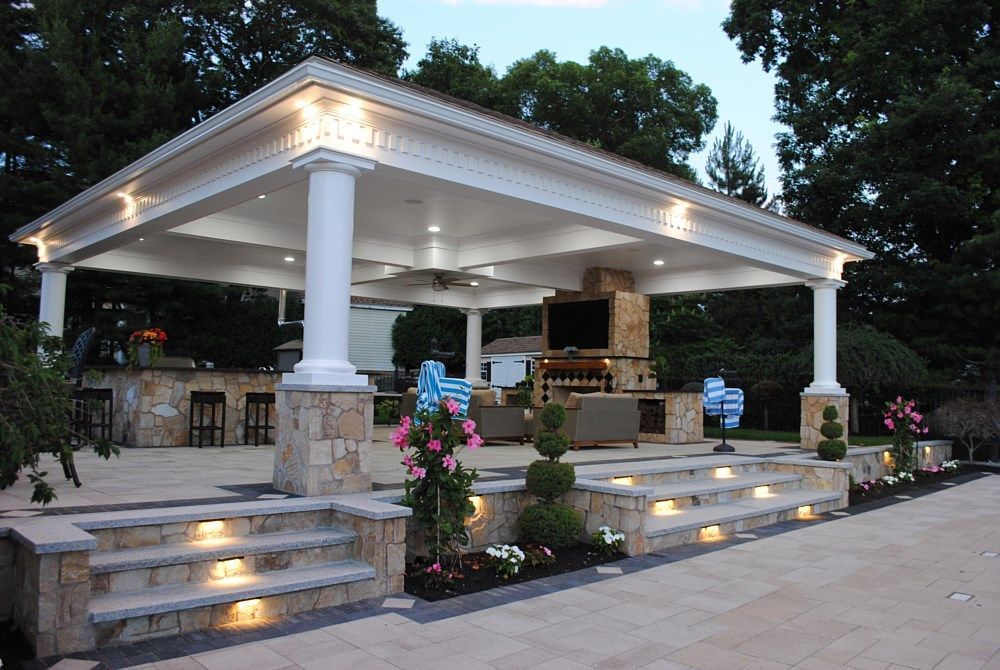 A covered outdoor patio with a stone fireplace, bar seating, and illuminated steps at twilight.