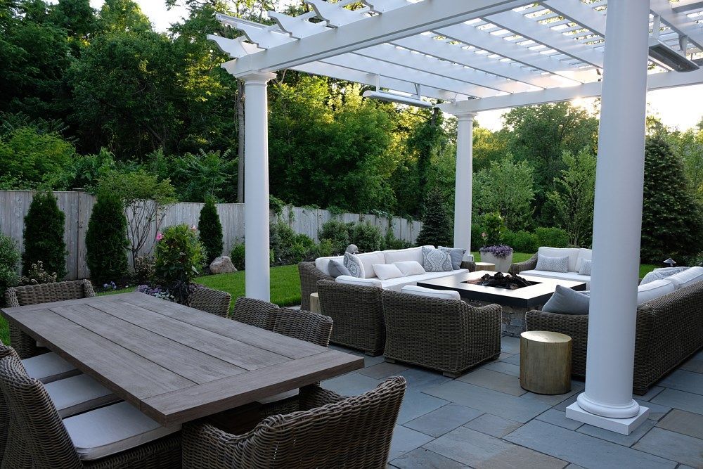 A patio featuring a large wooden dining table and a woven sectional sofa around a fire pit under a white pergola.