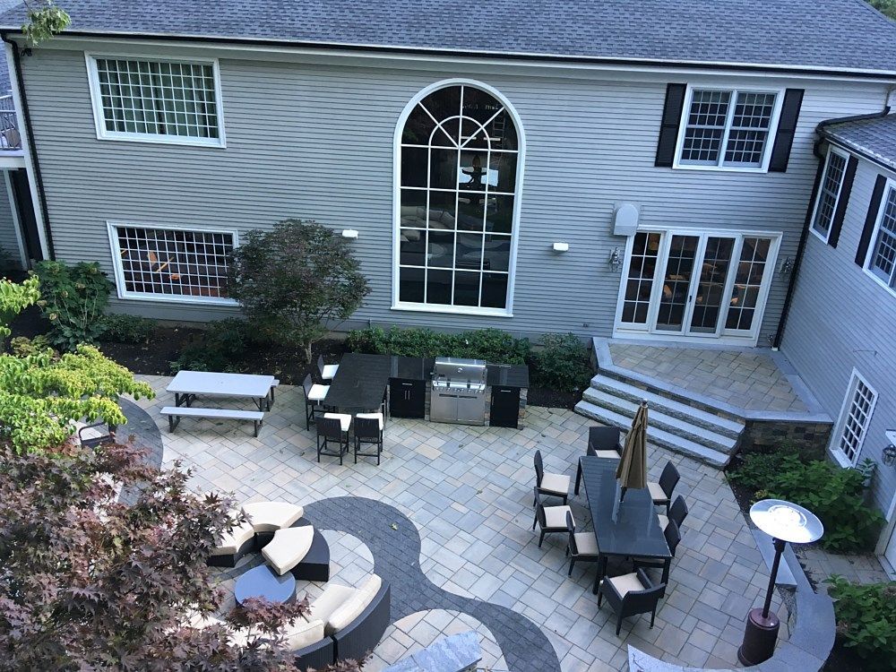 A high-angle view of a grey house with a stone patio featuring dining sets, lounge seating, and an outdoor grill.