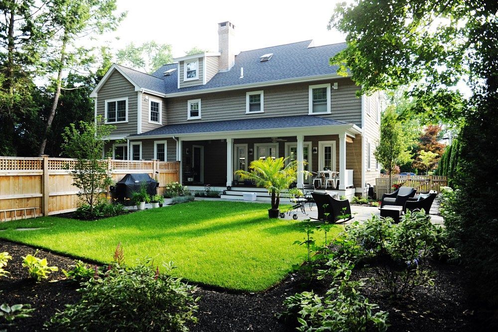 A two-story grey house with a manicured green lawn, a wooden fence, and a patio area with outdoor furniture.