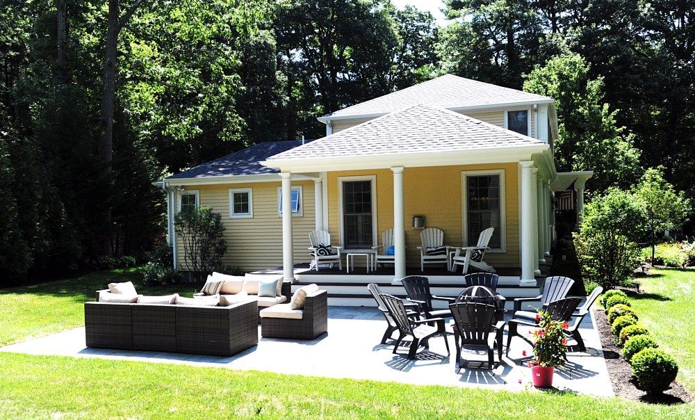 A yellow house with a covered porch and patio, featuring outdoor furniture, surrounded by a lawn and mature trees.