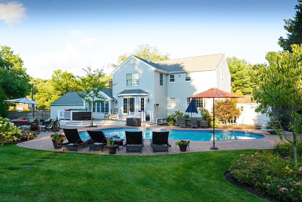 A sunny backyard with a light-colored two-story house, a swimming pool, lounge chairs, and an umbrella on a stone patio.