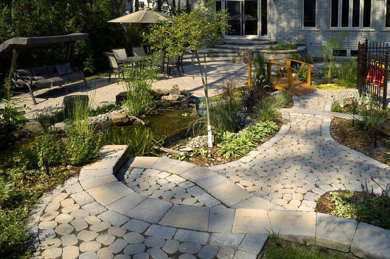 A stone-paved garden path leads toward a small pond, a wooden bridge, and a patio area with seating under an umbrella.