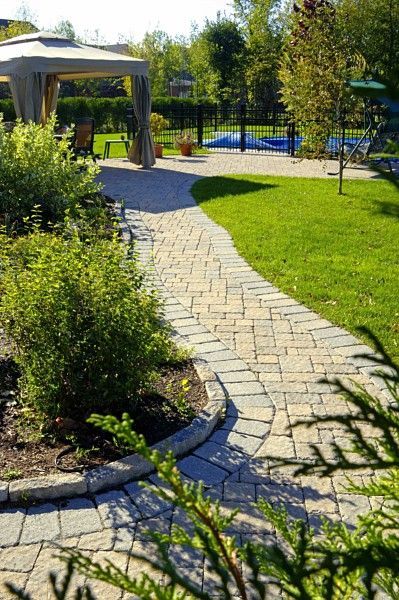 A stone paver pathway winds through a sunny backyard toward a gazebo and a fenced patio area.