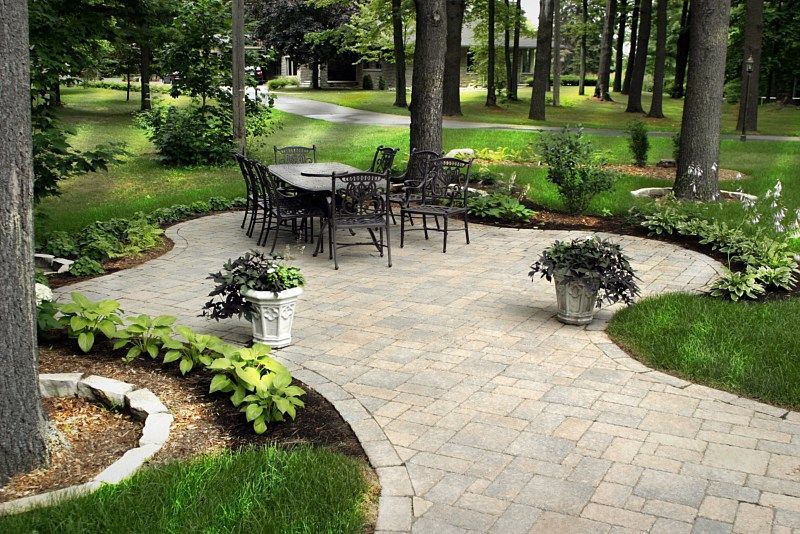 A stone patio with a metal dining set surrounded by lush trees, garden beds, and manicured lawn.