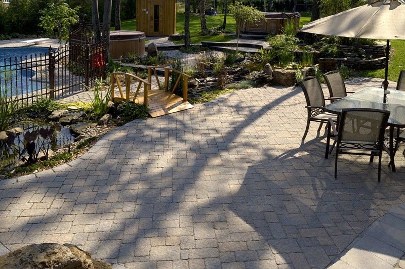 A sunny outdoor patio with paver stones, a dining set under an umbrella, a small wooden footbridge, and a pond.