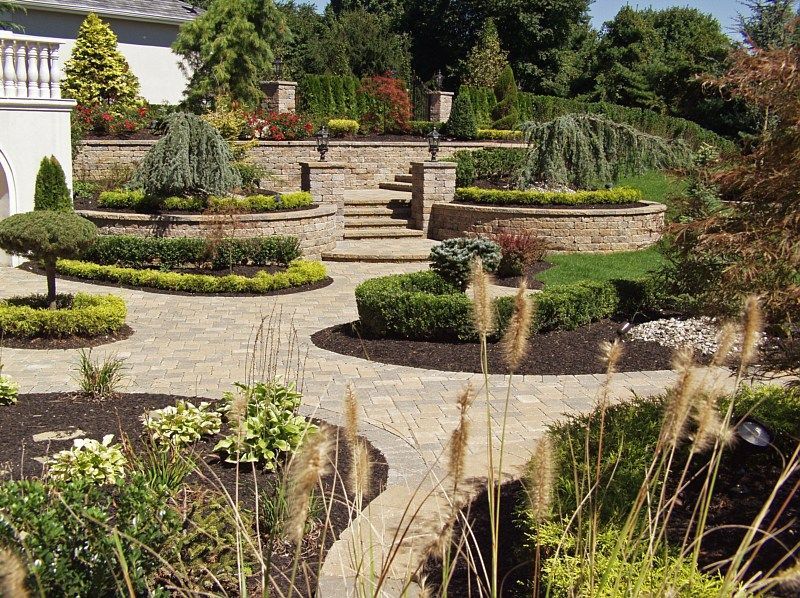 A landscaped garden with stone stairs, tiered retaining walls, manicured shrubs, and a paved patio area.