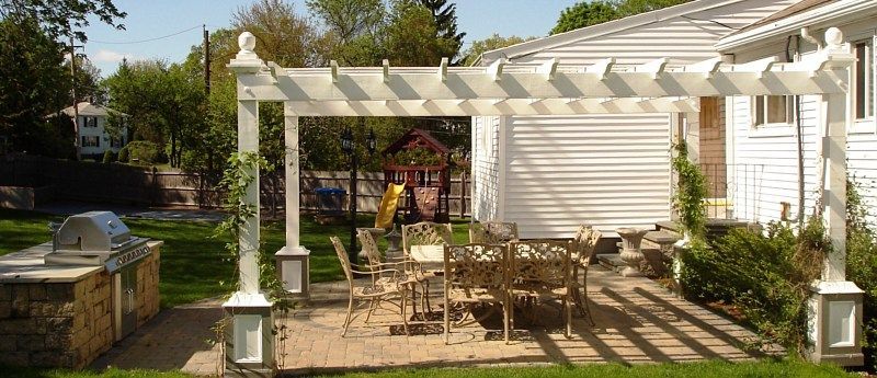 A backyard stone patio features a white pergola over a dining set, with a built-in outdoor grill on the left.