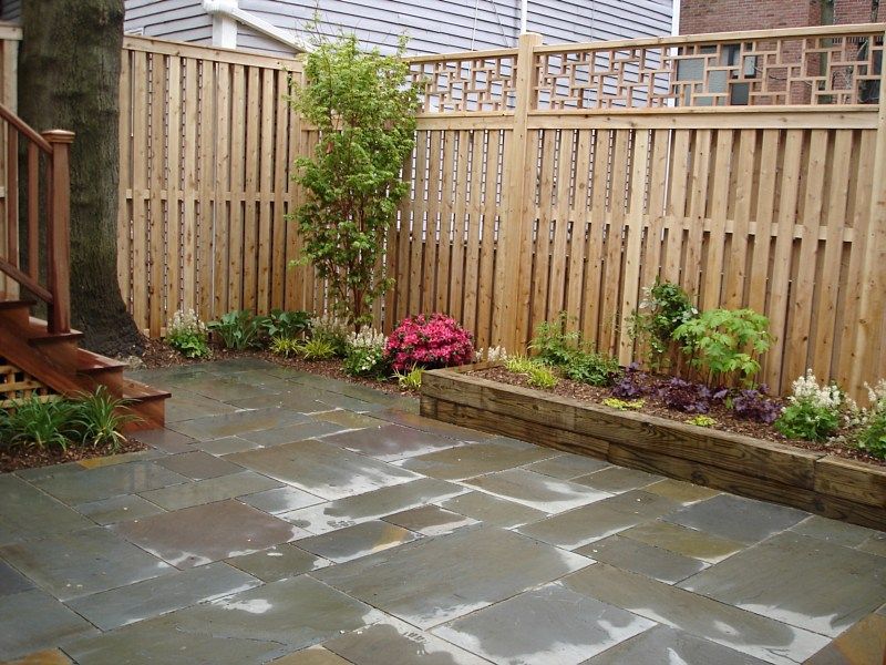 A stone patio with a wood fence, a raised garden bed with various plants, and stairs leading up on the left.