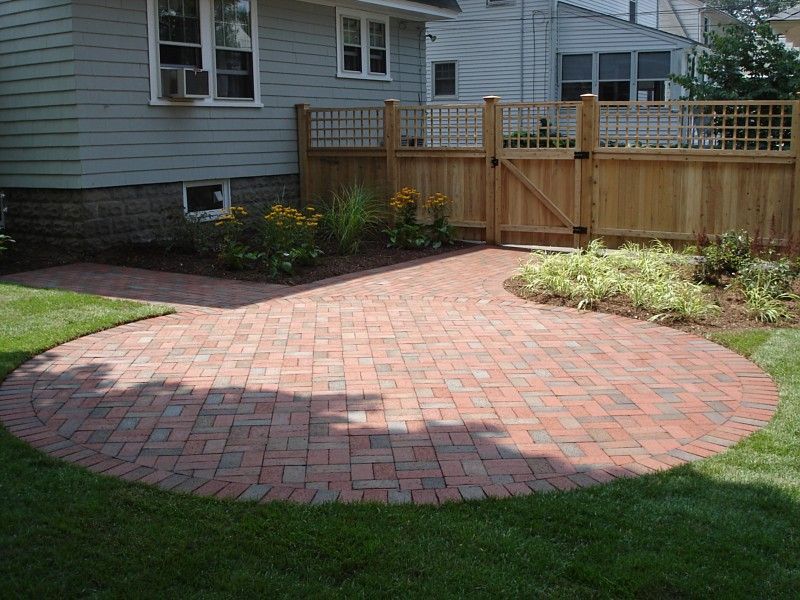 A circular red-brick patio in a backyard with a wooden privacy fence, garden beds, and green grass.
